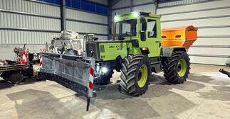 Ein in einer Halle stehender Traktor mit angebautem Salzstreuer und einem Schneepflug